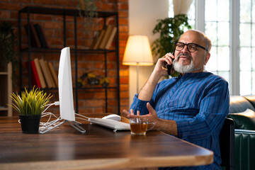 Indian elderly man in kurta works on desktop, talks on the phone, and drinks coffee at home