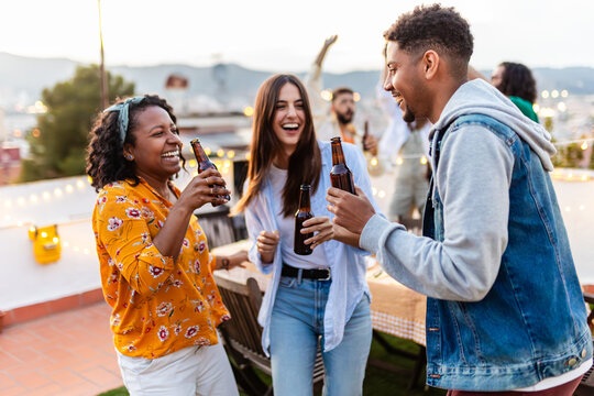 Young group of diverse friends enjoying rooftop summer party together. Millennial people having fun while dancing, drinking and feeling happy at night celebration on terrace