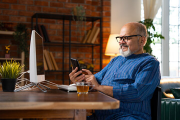 Indian elderly man in kurta works on desktop, talks on the phone, and drinks coffee at home