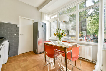 A sleek, modern kitchen featuring a wooden dining table, orange chairs, sunflowers in a vase, and large windows allowing natural light to fill the space.