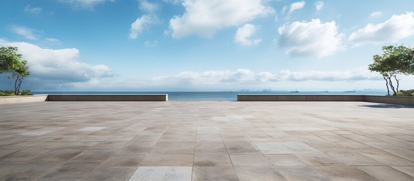 Empty beachfront park with vast empty stone floor, blue sky with clouds, calm sea in the background, flanked by green trees on either side.