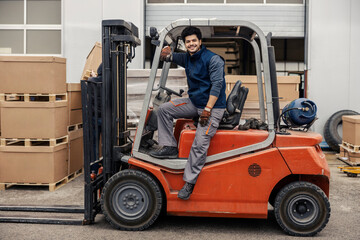 Full length of interracial forklift driver posing on forklift and smiling at camera at industrial zone.