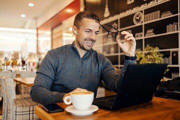 Young man taking off his eyeglasses while examining documentation on laptop from coffee shop.