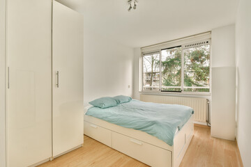 A bright, minimalist bedroom featuring a bed with light blue bedding, sleek storage furniture, and large windows that bring in natural light.