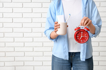Young woman with toothbrush, tube of paste and alarm clock on white brick background