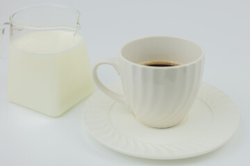 Close-up of a white mug with black coffee standing on a saucer, next to it is a jug of milk on a white background. The concept of invigorating drinks, coffee break, coffee with milk. High quality