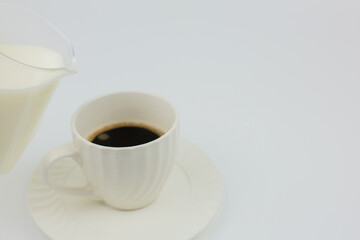 Close-up side view of a white mug with black coffee standing on a saucer, next to a jug of milk is brought to a mug on a white background with space for text. The concept of invigorating drinks