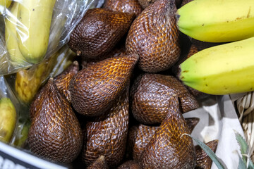A close-up of fresh salak fruits, also known as snake fruit, with scaly brown skin, displayed next to bananas in a market setting.