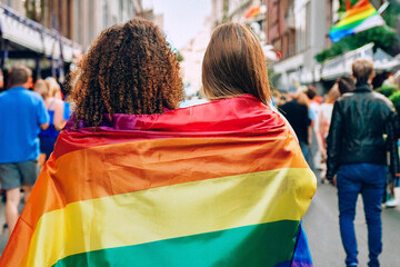 Two people wrapped in a rainbow flag at an LGBTQ+ pride parade, walking together in unity and love. The vibrant colors of the flag contrast with the lively urban background of diverse participants.