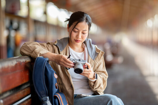 Smiling asian woman is looking through her camera while sitting on the wooden benchmark.