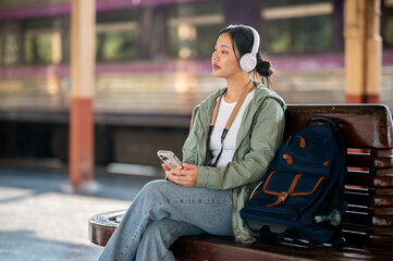 Asian woman is listening to music with headphones while sitting on wooden bench in the train station