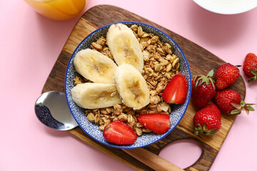 Wooden cutting board, bowl with tasty granola, banana and strawberries on pink background, closeup