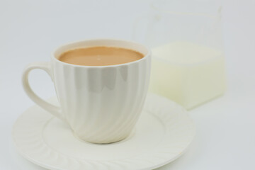 A close-up of a white mug with a saucer in which coffee with milk is poured stands on a white background, next to it a jug of milk is out of focus. Coffee with milk concept, coffee break. High quality