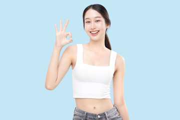 Beautiful young Asian woman with healthy and perfect skin shows OK signal on isolated blue...