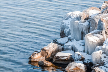 Big stones on the shore by the sea are covered with ice. Ice cliffs on the lake. Winter landscape with river and cliffs. Cold waters of a frozen lake