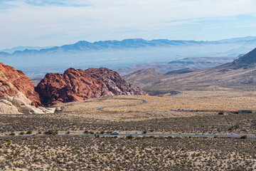 An expansive desert landscape featuring stunning red rock formations, contrasting with the soft earth tones of the surrounding terrain and distant mountains under a clear sky.