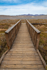 Fototapeta premium A picturesque wooden boardwalk stretches across a sunlit landscape, leading the viewer's eye toward distant mountains and showcasing the beauty of nature's simplicity.