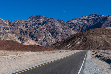 A remote desert road stretches towards towering mountains under a clear blue sky, illustrating the vastness of the landscape and the beauty of isolation.