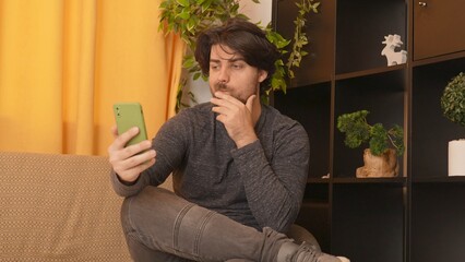 Adult man sitting on the sofa in the room holds smartphone watching social media, video content,...