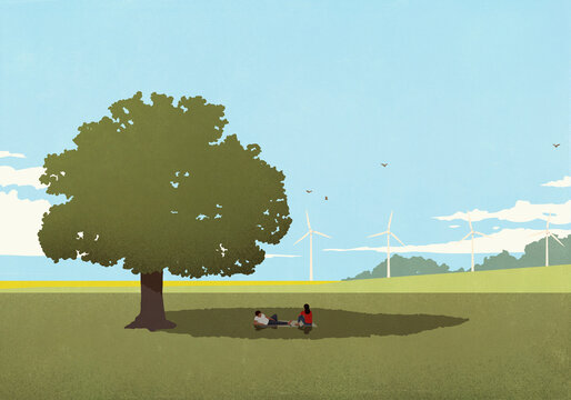 Couple relaxing on blanket in shade of tree in sunny field with wind turbines in the background 
