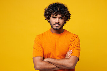 Young sad mad frowning suspicious Indian man he wearing orange t-shirt casual clothes hold hands crossed folded looking camera isolated on plain yellow background studio portrait. Lifestyle concept.