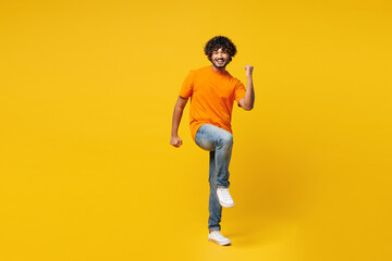 Full body young happy overjoyed cheerful cool Indian man he wear orange t-shirt casual clothes doing winner gesture celebrate clenching fists say yes raise up leg isolated on plain yellow background.