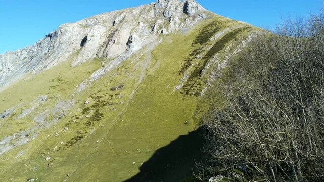 Impressive rocky mountain peak with green slopes and wooden shelter in mount txindoki, gipuzkoa. Hiker wearing blue jacket resting near wooden structure