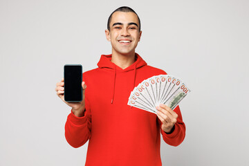Young middle eastern man wear red hoody casual clothes hold in hand fan of cash money in dollar banknotes use blank screen mobile cell phone isolated on plain solid white background Lifestyle concept