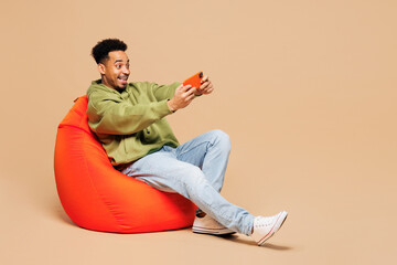 Full body young man of African American ethnicity wear green casual clothes sit in bag chair use...