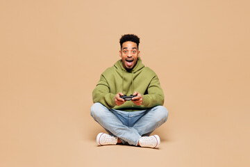 Full body young man of African American ethnicity wearing green hoodie casual clothes sit hold in hand play pc game with joystick console isolated on plain pastel beige background. Lifestyle concept. © ViDi Studio