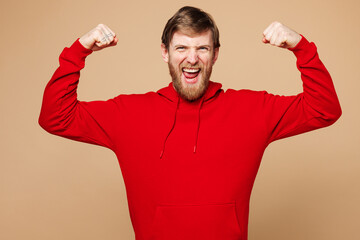 Young strong sporty fitness man he wear red hoodie casual clothes showing biceps muscles on hand demonstrating strength power isolated on pastel plain light beige background studio. Lifestyle concept.