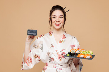 Young smiling happy woman wear kimono clothes eat raw fresh sushi roll served on black plate hold credit bank card look camera isolated on plain beige background studio Japanese food delivery concept
