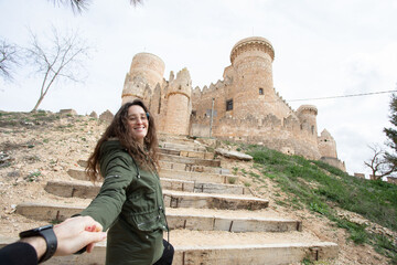 Una mujer con una chaqueta verde coge la mano de su pareja con un imponente castillo medieval de...