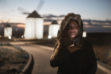 Fototapeta premium Una mujer delante de molinos de viento tradicionales de Castilla-La Mancha se alza en el paisaje al anochecer, evocando la historia y la cultura de la región inmortalizada en Don Quijote de la Mancha.