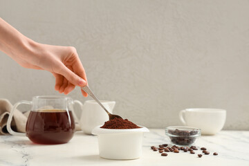 Female hand with bowl of coffee powder, spoon, beans, jugs and cup on light background