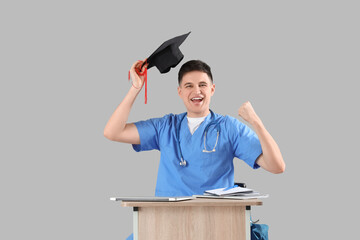 Happy medical intern with graduation hat at table on grey background
