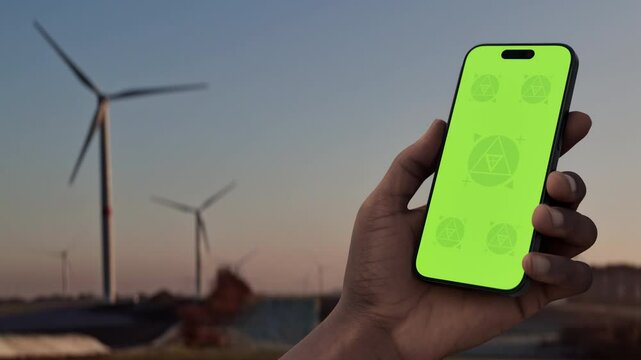 GREEN SCREEN CHROMA KEY: A video showcasing a phone in a man's hand, set against a rural wind farm backdrop. The video serves as a mockup for eco, energy, or environmental apps
