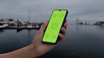 GREEN SCREEN CHROMA KEY Hand holding smartphone with green screen at harbor on an overcast day, ideal for content placement in a waterfront urban setting