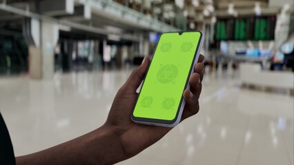 GREEN SCREEN CHROMA KEY A hand holding a smartphone with a green screen in an airport terminal. Blurred travelers and modern architecture in the background