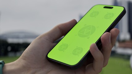 GREEN SCREEN CHROMA KEY Person holding a smartphone with a green screen outdoors, overlooking a modern urban skyline and grassy area on a cloudy day