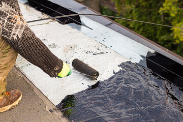 a young worker treats a roof with bitumen primer before applying rolled asphalt roofing using a...