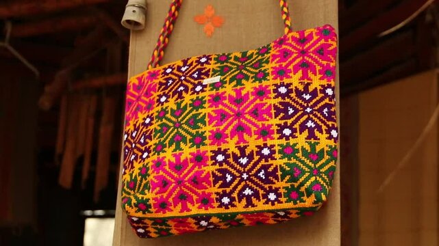 Colorful embroidered textile bag with geometric and floral patterns, orange and purple tones, fringed edges. 
