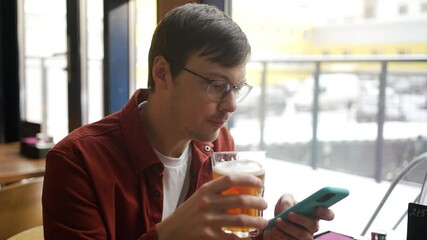 Casually dressed young man relaxing with beer, browsing smartphone while sitting at cafe window during winter day, enjoying leisure moment