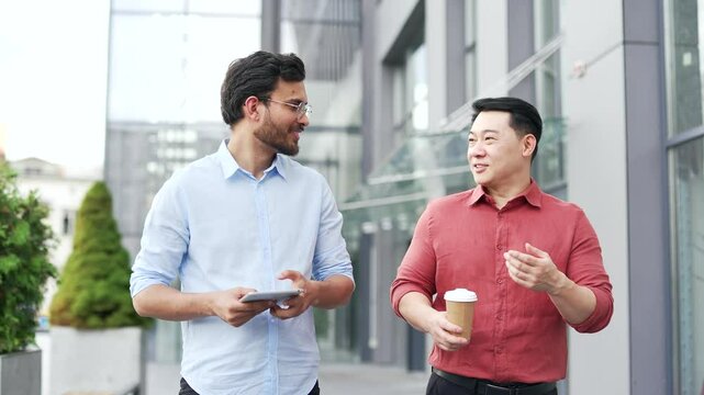 Two happy smiling businessmen engaging in discussion while walking outside modern office. One holds a tablet, another enjoys coffee. Teamwork and friendship among professionals in an urban setting