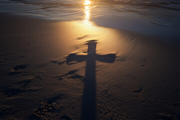 The shadow of a wooden cross stretches across the sand at sunset, illuminated by the golden glow of the setting sun, symbolizing faith, hope, and reflection in a serene coastal setting.