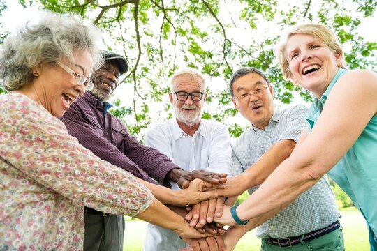 Diverse group of elderly people, men and women, smiling and stacking hands outdoors. Multicultural seniors enjoying teamwork and friendship in nature. Senior diverse people as a team.
