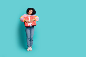 Happy young woman in stylish casual attire holding a red gift box against a teal background,...
