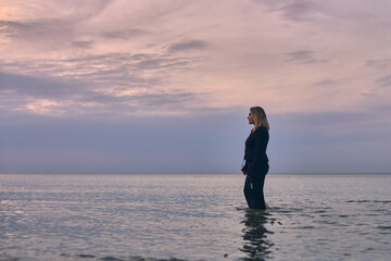 Young woman in black dress wades through calm ocean waters during tranquil sunset. Pink hues of sky evoke sense of solitude and peace, symbolizing introspection