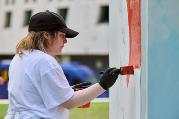 Young adult female artist, with light hair and casual attire, paints vivid mural. Outdoors setting...