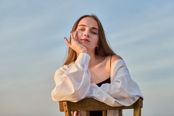 Young woman in white blouse sits on chair, gazing contemplatively into distance. Set against serene outdoor backdrop with soft lighting, mood is calm and introspective